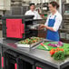 A woman in a blue apron opening a black Metro Mightylite food pan carrier with a red door to reveal a tray of salad.