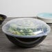 A black Fineline high profile plastic catering bowl filled with salad and covered with a clear lid.