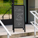 An Aarco black aluminum A-frame chalkboard sign with slate gray accents on the sidewalk with vegetables and prices.