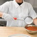 A person in a white coat using a Vollrath Orange Solid Round Spoodle to pour red sauce into a bowl.