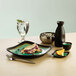 A black Libbey stoneware sake cup with a green liquid in it on a table with a plate of food.