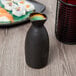 A black Libbey stoneware sake bottle filled with green liquid on a table with sushi.