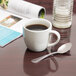 A Tuxton Milano white porcelain mug filled with coffee sits on a table next to a magazine and a spoon.