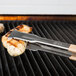 A pair of Vollrath VersaGrip tongs on meat cooking on a grill.