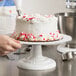 A person using an Ateco cake turntable to decorate a white cake with red and pink hearts.