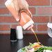 A hand holding a Tablecraft wide mouth squeeze bottle with orange liquid pouring onto a plate of salad.