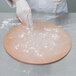 A person wearing gloves uses a pizza peel to spread flour on dough.