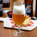 A Libbey stemmed pilsner glass filled with beer on a table next to a plate of fries and bottles.