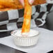 A person holding a french fry in a small clear fluted plastic ramekin of dipping sauce.