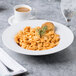 A white porcelain pasta bowl filled with pasta and a piece of bread on a table with a cup of coffee.
