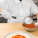 A person in a white shirt and gloves using a Vollrath Black Solid Round Spoodle to pour red sauce into a bowl.