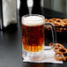 A customizable SAN plastic beer mug filled with beer with a handle on a table with pretzels.