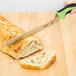 A Mercer Culinary Millennia Colors bread knife with a green handle cutting a piece of bread on a cutting board.