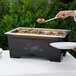 A person serving food from a Sterno Full Size ChalkBoard Chafer on a buffet table outdoors.
