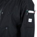 A person wearing a Mercer Culinary Millennia black long sleeve chef jacket with a phone and pen in the front pocket.