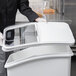 A person in a chef's uniform holding a clear plastic lid over a white Baker's Mark ingredient bin.
