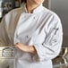 A woman wearing a Mercer Culinary white long sleeve chef jacket in a professional kitchen holds a metal colander.