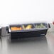 A San Jamar 3-compartment condiment bar with fruit and vegetables in it on a counter.
