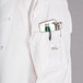 A Mercer Culinary white chef jacket with royal blue piping and pockets full of chef items.