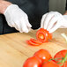 A person wearing white gloves using a Mercer Culinary Renaissance forged tomato knife to cut tomatoes on a table.
