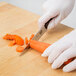 A hand in a white glove using a Victorinox serrated utility knife to cut a carrot on a wooden cutting board.