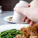 A hand holding a white bone china pepper shaker over a plate of food.