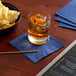 A glass of liquid with ice and a bowl of Choice navy blue cocktail napkins on a table.
