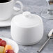A white Reserve by Libbey bone china sugar pot with a lid next to a bowl of fruit.