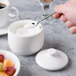 A hand holding a spoon over a white cup of sugar.