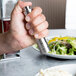 A hand holding a Fletchers' Mill stainless steel and acrylic salt mill over food.