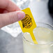 A hand using a yellow rectangular stirrer to stir a glass of lemonade with a yellow label.