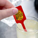A hand using a red rectangular stirrer in a glass of lemonade.