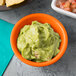An orange Tablecraft ramekin filled with guacamole on a table with a bowl of guacamole and salsa.