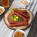 A Tablecraft acacia wood round serving board with food, sausages, pretzels, and peanuts on it.
