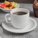 A cup of coffee sits on a Libbey Porcelana double well saucer next to a bowl of fruit.