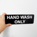 A hand holding a black and white Vollrath Traex sign that says "Hand Wash Only"