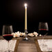 A lit ivory Hyoola taper candle on a table set with wine glasses and flowers.