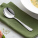 An Acopa stainless steel bouillon spoon on a green napkin next to a bowl of soup.