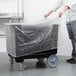 A man in a chef's uniform holding a plastic bag over a Cambro black dish caddy with plastic wrap over it.