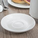 A Libbey bright white porcelain saucer on a wood surface with a white cup on it.
