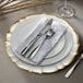 A white glass charger plate with a scalloped edge and gold trim with silverware on it.