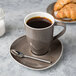 A Schonwald dark gray porcelain saucer with a cup of coffee and a spoon on it, next to a plate of croissant.