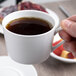 A hand holding a Schonwald Continental white porcelain cup of coffee over a plate of food.