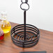 A black metal wrought iron condiment caddy on a wood surface with a glass jar and a bottle inside.