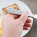 A hand holding a Libbey Cortland stainless steel bread and butter knife over a plate of food.