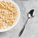A bowl of cereal with milk and a Libbey stainless steel teaspoon on a table.