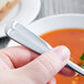 A hand holding a World Tableware Columbus stainless steel bouillon spoon over a bowl of soup.
