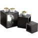 Three black wooden cube risers with bowls of food on top.