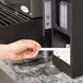A hand using a Vollrath black countertop lid organizer with a straw pocket to dispense a white stick.
