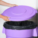 A hand holding a Carlisle purple flat round trash can lid over a purple trash can.